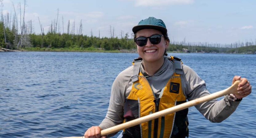 A person wearing a lifejacket and holding a paddle is sitting in a canoe and smiles for the photo.